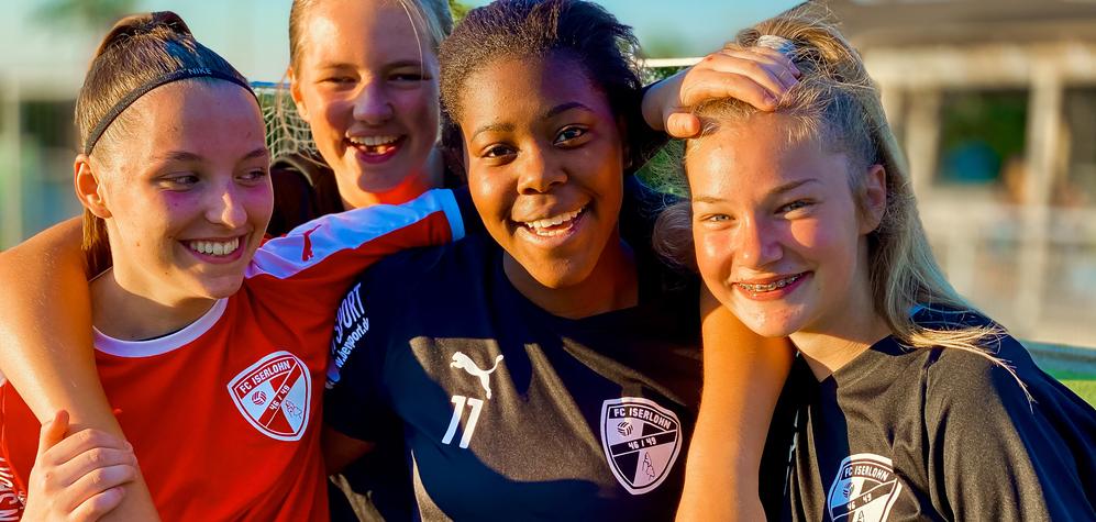 Vier junge Fußballspielerinnen in Teamtrikots posieren lachend gemeinsam auf dem Spielfeld.