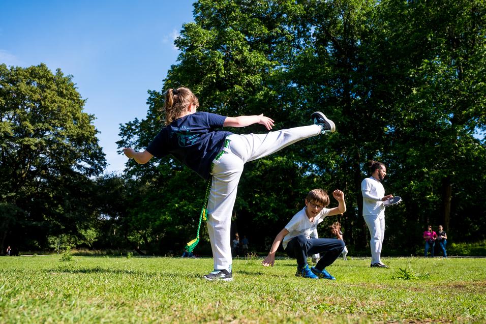 Zwei Kinder spielen im Park; eines tritt mit hohem Bein, das andere kniet und schaut überrascht. Grüne Bäume im Hintergrund.