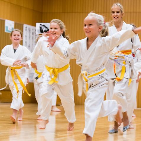 Gruppe von Kindern in weißen Karateanzügen mit gelben Gürteln, die fröhlich in einer Sporthalle herumrennen.