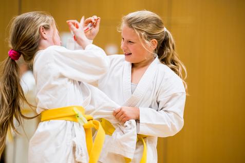 Zwei Mädchen in weißen Karateanzügen mit gelben Gurten üben freundschaftlich im Trainingsraum miteinander.