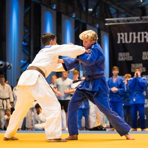 Zwei Jugendliche im Judoanzug kämpfen auf einer gelben Matte bei den Ruhr Games, während Zuschauer im Hintergrund beobachten.