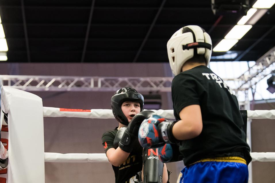 Zwei Kinder im Kickboxring, eines hebt den Fuß zu einem Kick, das andere bereitet sich auf einen Angriff vor.