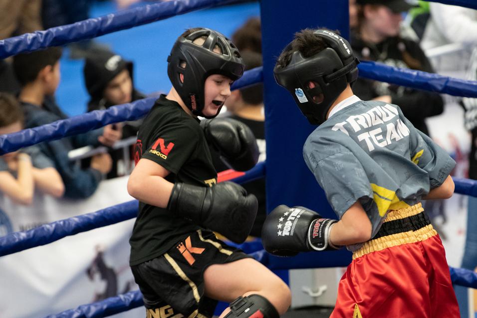 Zwei Kinder im Boxring, ausgerüstet mit Schutzausrüstung, in einem intensiven Kampfmoment während eines Wettkampfs.