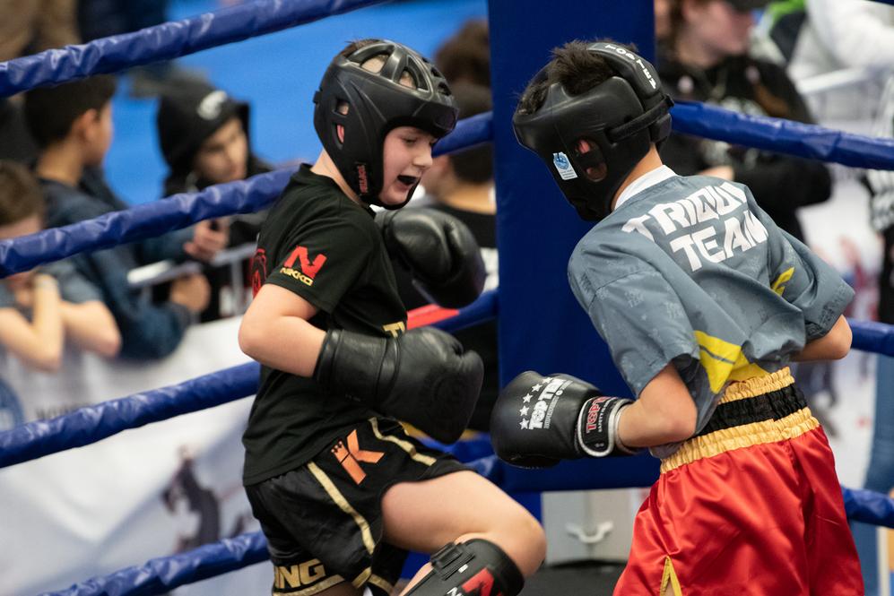 Zwei Kinder im Boxring, ausgerüstet mit Schutzausrüstung, in einem intensiven Kampfmoment während eines Wettkampfs.