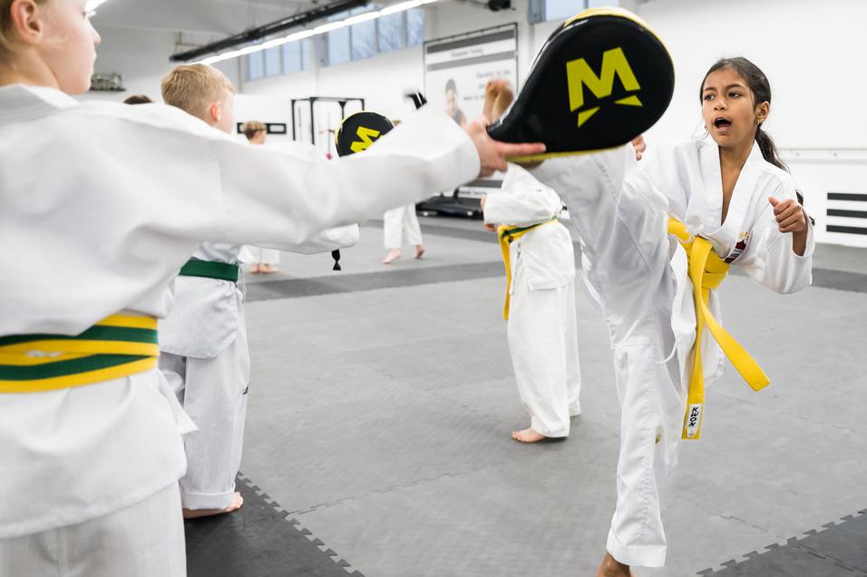 Kinder trainieren in weißen Karateanzügen, während ein Mädchen mit gelbem Gürtel einen hohen Kick ausführt.