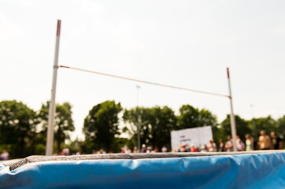 Nahaufnahme einer blauen Sprungmatte mit einer Hochsprunganlage und unscharfem Publikum im Hintergrund.