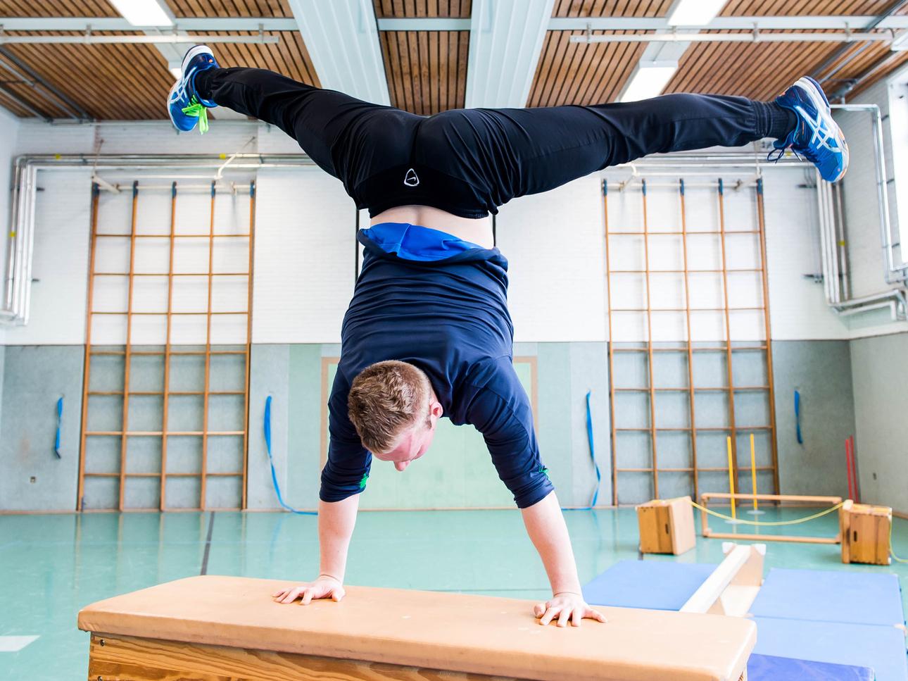 Junge macht Handstand auf einem Holztisch in einer Sporthalle mit verschiedenen Turngeräten im Hintergrund.