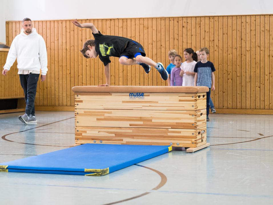 Ein Junge springt über ein Turngerät, während eine Gruppe Kinder und ein Trainer zuschauen. Sporthalle mit Holzverkleidung.
