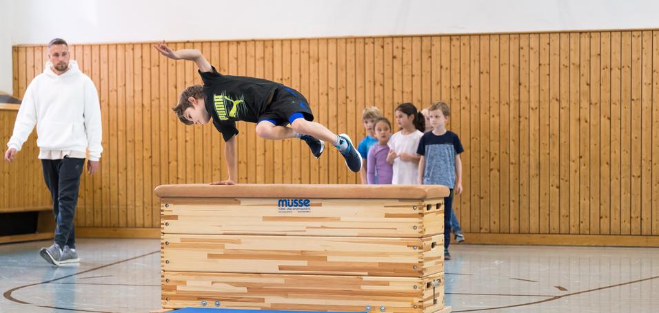 Ein Junge springt über ein Turngerät, während eine Gruppe Kinder und ein Trainer zuschauen. Sporthalle mit Holzverkleidung.