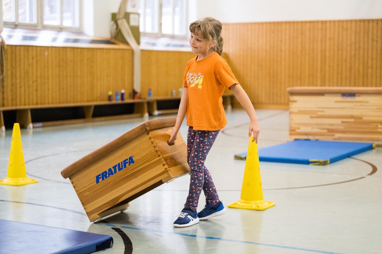 Mädchen in orangem T-Shirt trägt einen Turnkasten in einer Turnhalle, umgeben von gelben Hütchen und Matten.