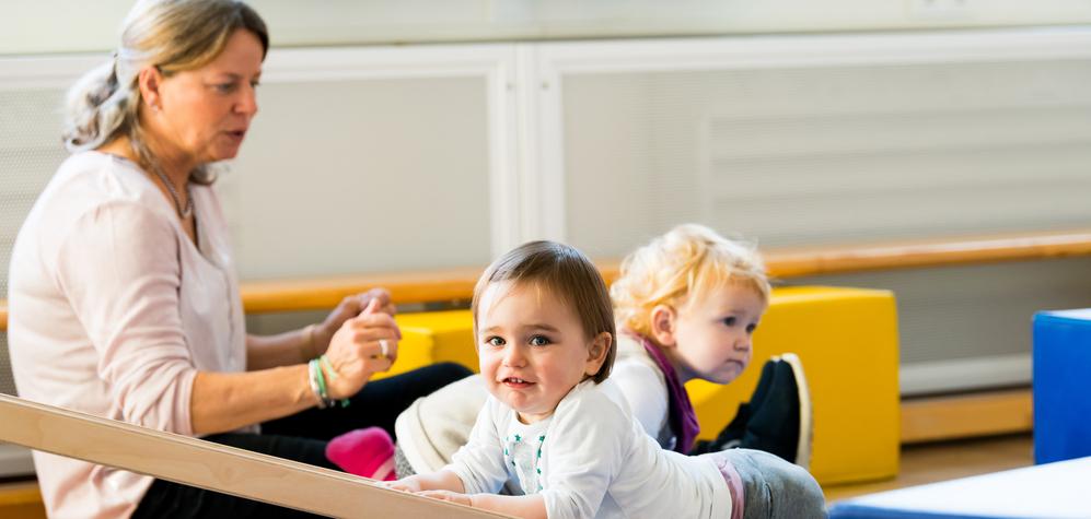 Zwei Kleinkinder spielen auf einer blauen Matte, während eine betreuende Person im Hintergrund sitzt.