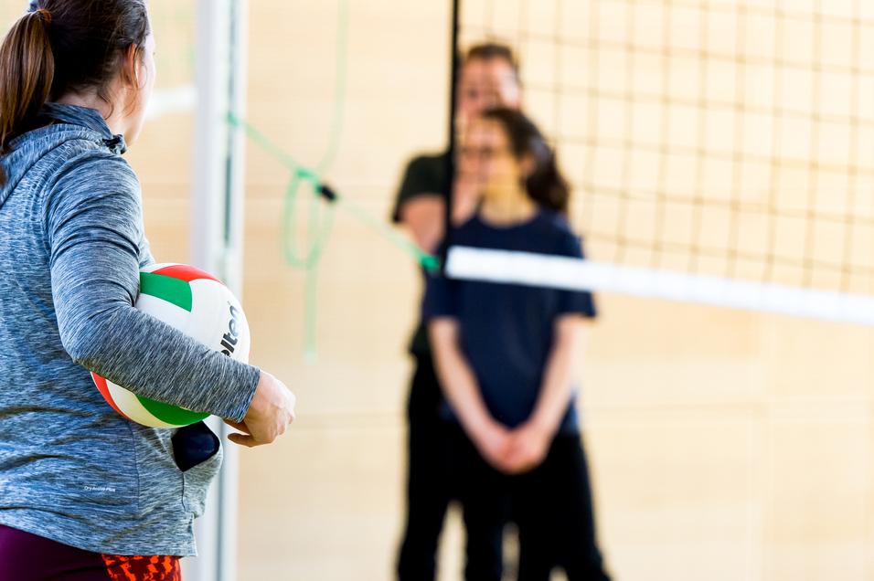 Eine Person mit einem Volleyball steht vor dem Netz, während zwei weitere Personen im Hintergrund warten.